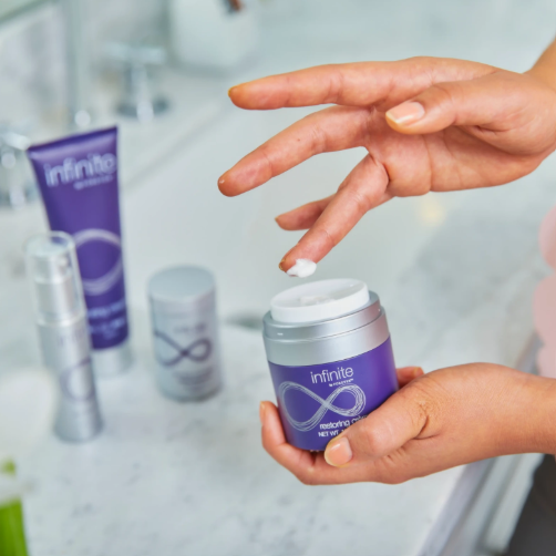 Crème restauratrice anti-âge Infinite Forever appliquée du bout des doigts, avec les autres produits de la gamme en arrière-plan