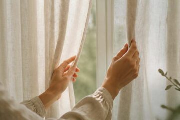 Mains ouvrant un rideau en lin pour laisser entrer la lumière du matin, un geste simple contre la fatigue de rentrée
